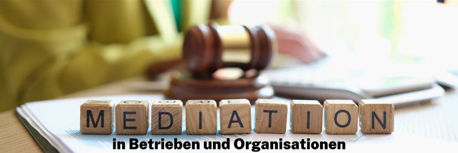 Holzwürfel mit dem Schriftzug „Mediation“, ein Richterhammer und eine geöffnete Akte im Hintergrund.